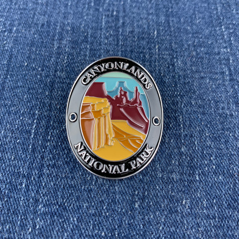 An oval enamel Walking Stick Medallion featuring a Canyonlands National Park landscape with red rock canyons and a yellow desert foreground, bordered by a silver-toned ring with white lettering, placed on a blue denim background.