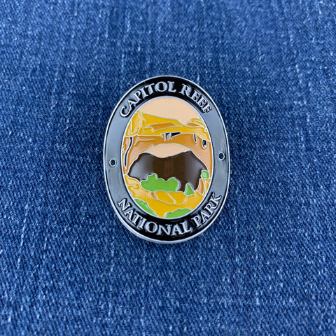 An oval enamel Walking Stick Medallion featuring a landscape illustration of Capitol Reef National Park, displayed on a blue denim fabric background.