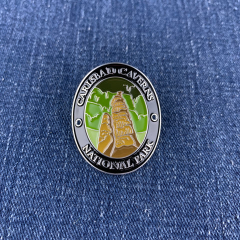 A circular enamel Walking Stick Medallion of Carlsbad Caverns National Park, featuring a gold-toned stalactite and green cave background, set against a blue denim fabric background.