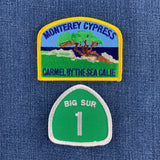 Two embroidered patches displayed on blue denim fabric: a rectangular 'Monterey Cypress - Carmel by the Sea Calif.' patch with a yellow border and scenic illustration, and a green shield-shaped 'Big Sur 1' patch below it.