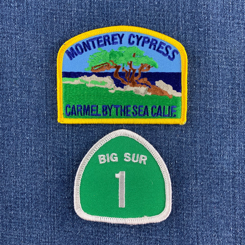 Two embroidered patches displayed on blue denim fabric: a rectangular 'Monterey Cypress - Carmel by the Sea Calif.' patch with a yellow border and scenic illustration, and a green shield-shaped 'Big Sur 1' patch below it.
