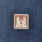 An embroidered square patch featuring a white silhouette of a Gothic cathedral on a brown background with a white stitched border displayed on a denim background