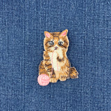 A small embroidered cat patch with a pink ball of yarn, placed on a blue denim fabric background.