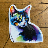A die-cut sticker in the shape of a cat's head and chest, featuring an abstract watercolor portrait of a tabby cat. The cat's fur is depicted in vibrant, saturated colors including blue, purple, pink, and yellow. The sticker is placed on a wooden surface.
