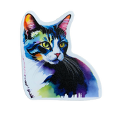 A die-cut sticker in the shape of a cat's head and chest, featuring an abstract watercolor portrait of a tabby cat. The cat's fur is depicted in vibrant, saturated colors including blue, purple, pink, and yellow. The sticker is placed on a wooden surface.