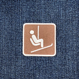 A square brown embroidered patch with a white border and a white ski lift icon, placed on a blue denim fabric background.