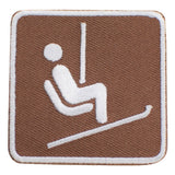 An embroidered square patch featuring a white icon of a seated person on a ski lift against a brown background, with a white stitched border.