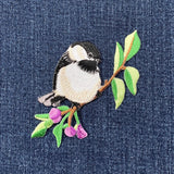 Embroidered patch of a bird on a branch with leaves and flowers on denim fabric.