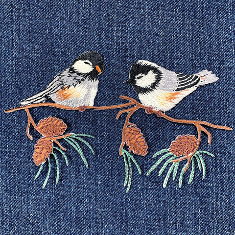 Embroidered design of two birds on a branch with pine cones on denim fabric.