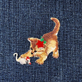 Embroidered patch of a cat playing with a mouse, wearing a red poinsettia flower on its head, on a denim background.