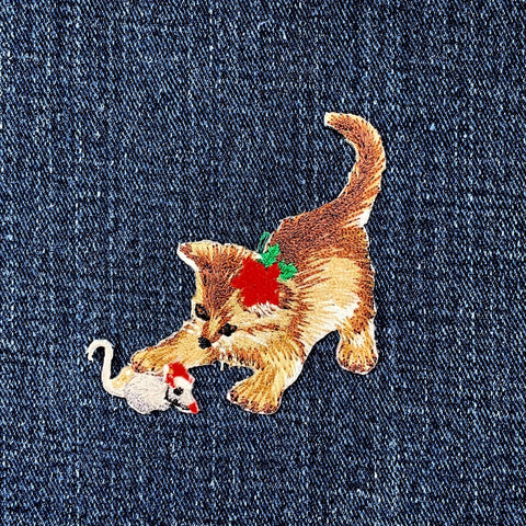 Embroidered patch of a cat playing with a mouse, wearing a red poinsettia flower on its head, on a denim background.