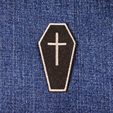 A coffin-shaped embroidered patch with a silver cross on a black background, shown on a blue denim fabric surface.