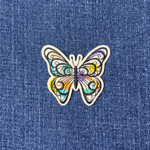 A colorful embroidered butterfly applique patch with antennae and wings on a denim background