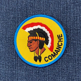 A round patch featuring an illustration of a Native American figure with a headdress, predominantly yellow with red and black accents, and the word 'COMANCHE' below on a denim background