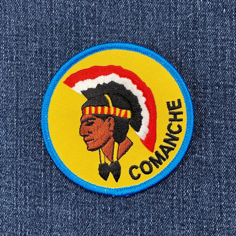 A round patch featuring an illustration of a Native American figure with a headdress, predominantly yellow with red and black accents, and the word 'COMANCHE' below on a denim background