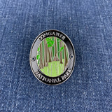 A circular metal Walking Stick Medallion featuring an illustration of Congaree National Park with green trees and a light green ground, bordered by a black ring with silver lettering, placed on a blue denim background.