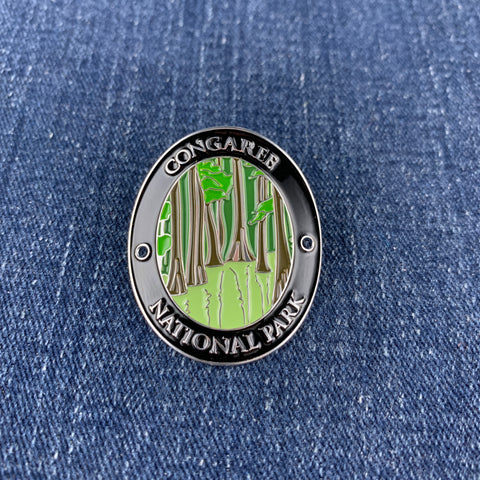 A circular metal Walking Stick Medallion featuring an illustration of Congaree National Park with green trees and a light green ground, bordered by a black ring with silver lettering, placed on a blue denim background.