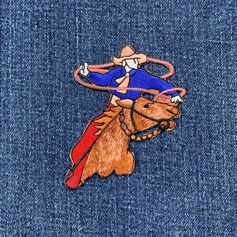 Embroidered patch of a cowboy on a horse with a lasso on blue denim fabric.