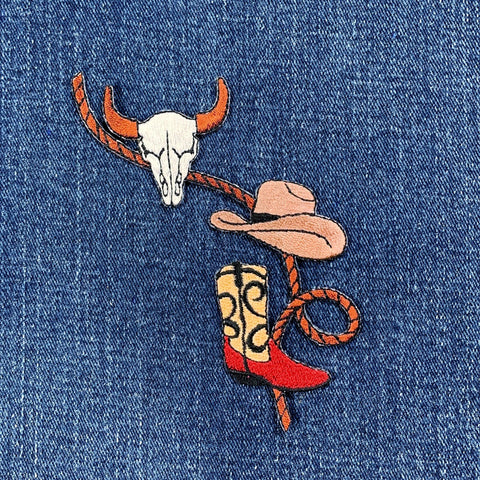 Embroidered patch on denim fabric featuring a cowboy hat, boot, and bull skull with a lasso.