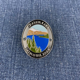 An oval metal enamel Walking Stick Medallion featuring a scenic landscape of Crater Lake National Park, displayed on a blue denim fabric background.