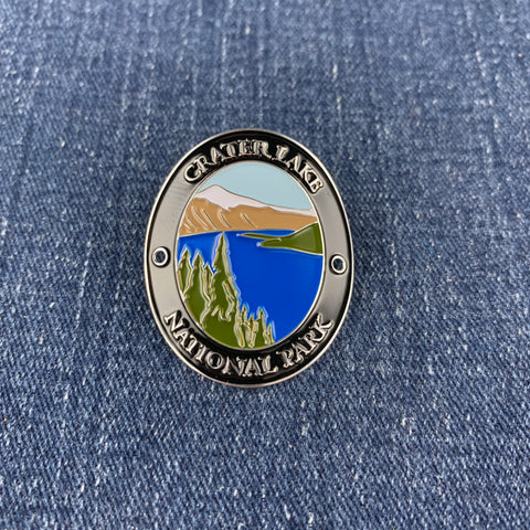 An oval metal enamel Walking Stick Medallion featuring a scenic landscape of Crater Lake National Park, displayed on a blue denim fabric background.