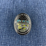 A circular enamel Walking Stick Medallion depicting Craters of the Moon National Monument, featuring a silver-toned border and landscape details in black, gray, teal, and lime green, placed on a blue denim background.