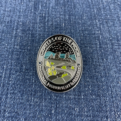 A circular enamel Walking Stick Medallion depicting Craters of the Moon National Monument, featuring a silver-toned border and landscape details in black, gray, teal, and lime green, placed on a blue denim background.