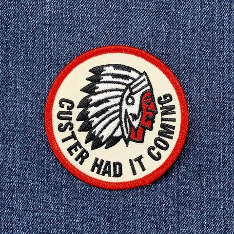 round patch with a red border showing a profile illustration of a Native American chief in a feathered war bonnet. Text reads: "CUSTER HAD IT COMING."