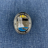 An oval metal enamel Walking Stick Medallion featuring a scenic depiction of Cuyuhoga Valley National Park, including waterfalls, mountains, and blue water, with the text 'CUYAHOGA VALLEY NATIONAL PARK' around the border, shown pinned on a blue denim background.