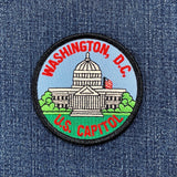 Circular embroidered patch depicting the United States Capitol Building with its white dome, set against a light blue sky and surrounded by green grass. The text 'WASHINGTON, D.C.' is stitched in red above, and 'U.S. CAPITOL' is stitched in red below, all on a blue denim background