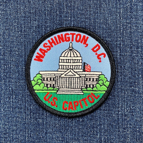 Circular embroidered patch depicting the United States Capitol Building with its white dome, set against a light blue sky and surrounded by green grass. The text 'WASHINGTON, D.C.' is stitched in red above, and 'U.S. CAPITOL' is stitched in red below, all on a blue denim background