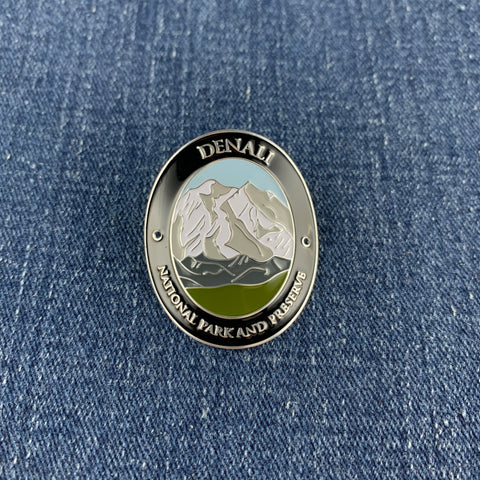 An oval metal enamel medallion featuring a mountain landscape with the text 'DENALI NATIONAL PARK AND PRESERVE' on a blue denim background.