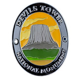 An oval enamel Walking Stick Medallion featuring a scenic depiction of Devils Tower National Monument, with a gray tower, green and yellow landscape, and a black outer border with silver lettering.