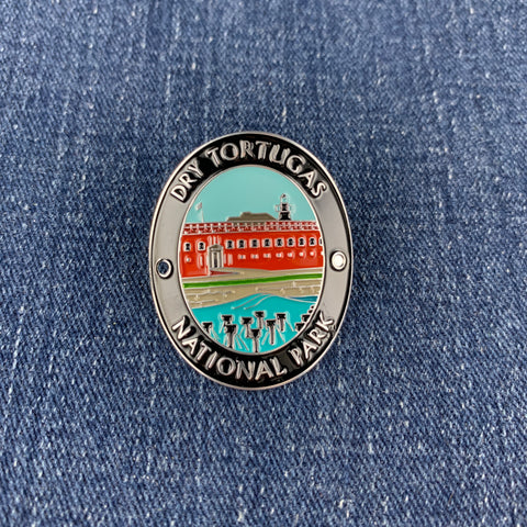 An oval enamel Walking Stick Medallion featuring a colorful illustration of Dry Tortugas National Park, including a red building, lighthouse, and turquoise water, with the text 'DRY TORTUGAS NATIONAL PARK' on a blue denim background.