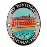 An oval Walking Stick Medallion for Dry Tortugas National Park, featuring an enamel illustration of the red Fort McHenry with a lighthouse, a turquoise sky and bay, and silver-toned lettering on a black background.