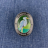 An oval enamel Walking Stick Medallion featuring the Everglades National Park logo with a blue heron, set against a blue denim fabric background.