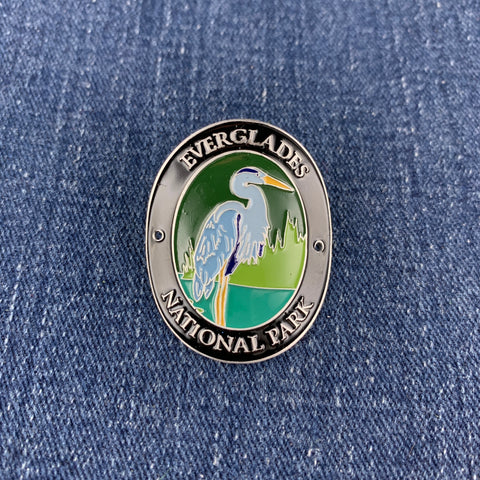 An oval enamel Walking Stick Medallion featuring the Everglades National Park logo with a blue heron, set against a blue denim fabric background.