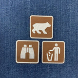 Three square brown embroidered patches with white icons are arranged on a blue denim background. The top patch shows a bear, the bottom left shows binoculars, and the bottom right shows a person throwing trash.