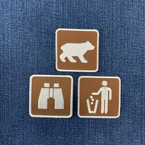 Three square brown embroidered patches with white icons are arranged on a blue denim background. The top patch shows a bear, the bottom left shows binoculars, and the bottom right shows a person throwing trash.