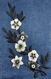 Embroidered appliqué featuring a winding vine of six flowers and several leaves, stitched in white, black, and grayscale thread, against a background of bright blue denim fabric.