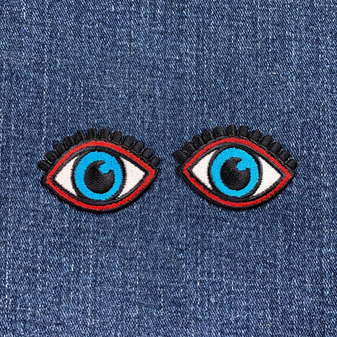 two patches depicting a pair of blue eyes with black eyelashes and an iron on backing, arranged side by side on a denim background