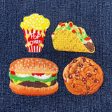 Set of four food-themed patches on denim fabric: pop corn, taco, hamburger, and cookie.