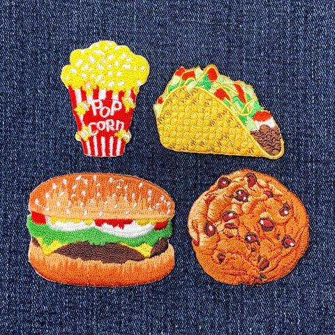 Set of four food-themed patches on denim fabric: pop corn, taco, hamburger, and cookie.
