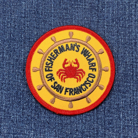 Round yellow patch with a red border depicting a red crab inside the spokes of a brown ship's wheel. Text around the wheel reads "FISHERMAN'S WHARF OF SAN FRANCISCO."