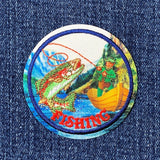 A circular embroidered patch featuring a fisherman casting a line toward a large fish, with the word 'FISHING' in red at the bottom, shown on a blue denim fabric.