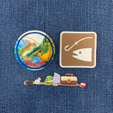 Three fishing-themed patches arranged on a blue denim fabric background: a round pin with a colorful fish and the word “FISHING,” a square tan pin with a white fishing hook and fish silhouette, and a long pin depicting a fishing rod, frog, tackle box, and a red-and-white striped ball.