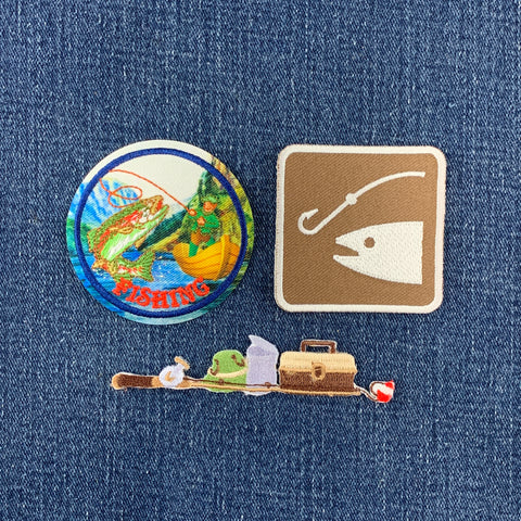Three fishing-themed patches arranged on a blue denim fabric background: a round pin with a colorful fish and the word “FISHING,” a square tan pin with a white fishing hook and fish silhouette, and a long pin depicting a fishing rod, frog, tackle box, and a red-and-white striped ball.