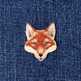 Embroidered patch in the shape of a fox's head with brown, white, and beige colors representing the fur and facial features on a denim background.