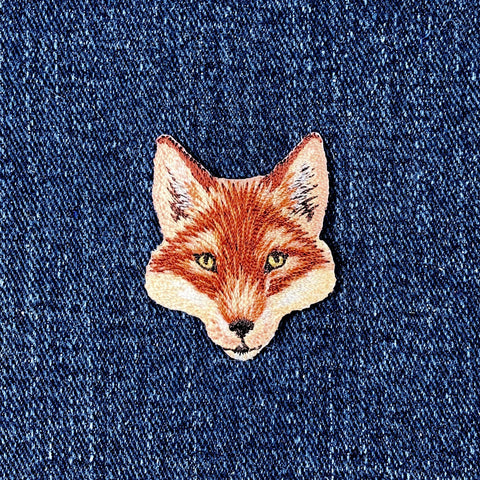 Embroidered patch in the shape of a fox's head with brown, white, and beige colors representing the fur and facial features on a denim background.