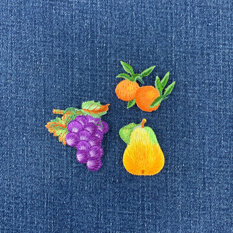 Three embroidered fruit patches on blue denim fabric: a purple grape cluster with green and orange leaves, two orange fruits with green leaves, and a yellow-to-green pear.